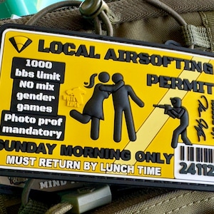 Può includere: Un cerotto giallo e nero con il testo "LOCAL AIRSOFTING PERMIT" e un codice a barre. Il cerotto ha una grafica di due figure stilizzate e una figura stilizzata che tiene una pistola. Il testo "SUNDAY MORNING ONLY MUST RETURN BY LUNCH TIME" è anche sul cerotto.