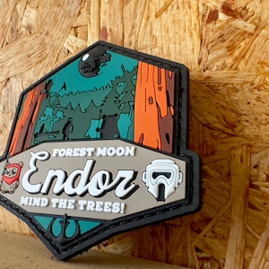 Può includere: Toppa esagonale con una scena della foresta e la scritta "FOREST MOON ENDOR MIND THE TREES!". Il design include Ewoks dei cartoni animati, alberi e un casco da stormtrooper. La toppa ha un bordo nero ed è ambientata su uno sfondo di venature del legno.