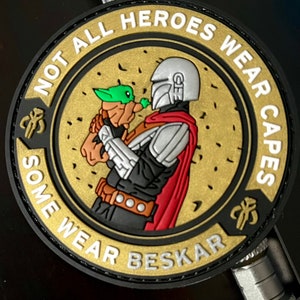 Puede incluir: Un parche redondo con una figura mandaloriana sosteniendo a Grogu. El parche tiene un fondo dorado con el texto "NOT ALL HEROES WEAR CAPES SOME WEAR BESKAR" en blanco. El mandaloriano lleva una armadura plateada y una capa roja.
