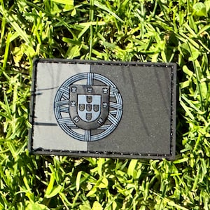 May include: A black and white embroidered patch featuring the Portuguese coat of arms. The patch is surrounded by a black border and has a textured fabric.
