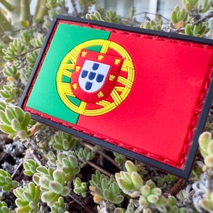 May include: A green and red embroidered patch with the Portuguese flag. The flag features a shield with a white background and seven blue five-pointed stars. The shield is surrounded by a yellow circle with a red border.