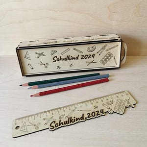 May include: A wooden pencil box with a school theme design. The box is engraved with the text "Schulkind 2024" and features various school supplies like pencils, rulers, scissors, and a compass. The box is accompanied by three colored pencils and a wooden ruler with the same design and text.