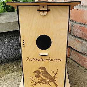May include: A wooden birdhouse with black trim and a hinged roof. The birdhouse has a round entrance hole and a small perch. The front of the birdhouse is decorated with a laser-engraved image of two birds on a branch and the text "Zwitscherkasten".