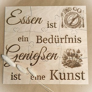 Könnte beinhalten: Ein Holzpuzzle mit dem Text "Essen ist ein Bedürfnis Genießen ist eine Kunst" (Essen ist ein Bedürfnis, Genießen ist eine Kunst). Das Puzzle ist aus hellem Holz gefertigt und hat eine natürliche Oberfläche. Der Text ist in das Holz graviert und leicht zu lesen.