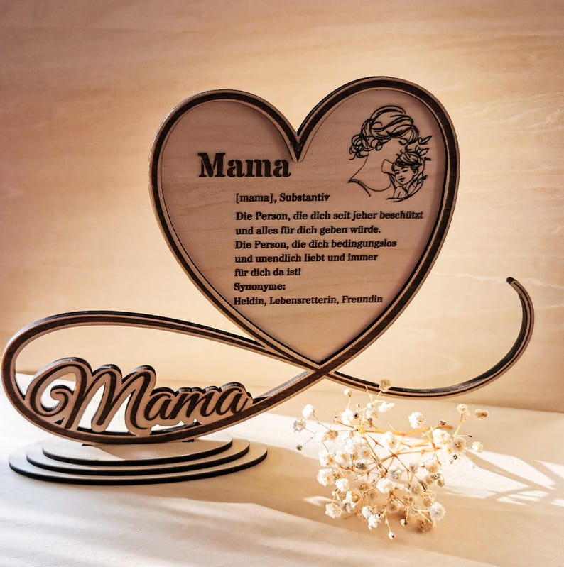 May include: Wooden heart-shaped sign with the word "Mama" in a decorative font. The sign also includes a definition of the word "Mama" in German, describing it as the person who protects and loves unconditionally. The sign is on a wooden base with an infinity symbol.