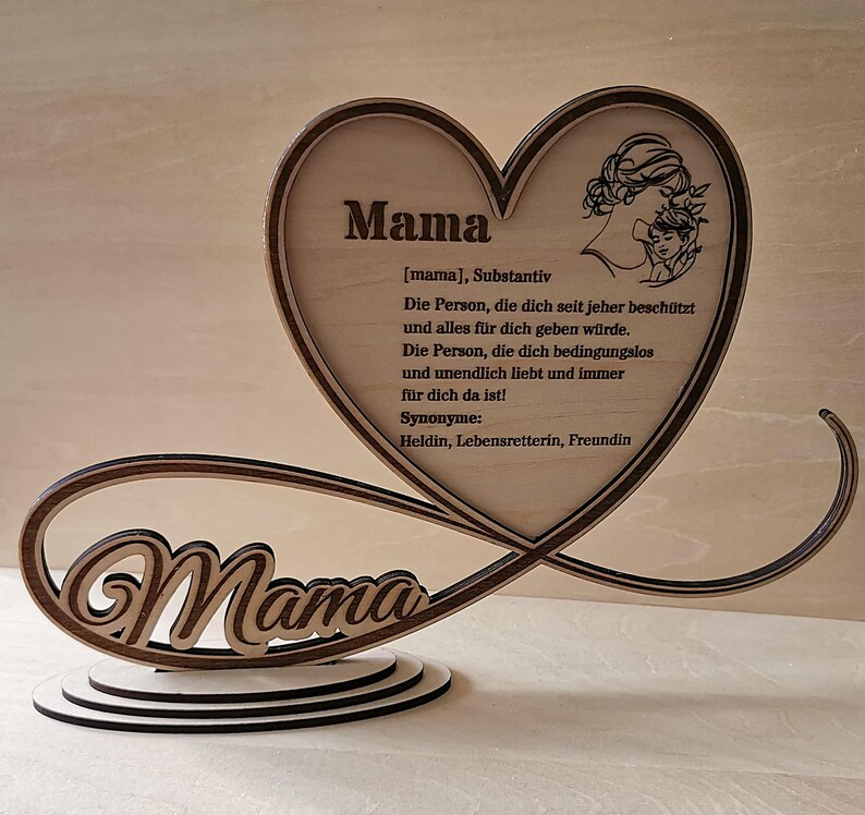 May include: Wooden heart-shaped sign with the word "Mama" in a decorative font. The sign also includes a definition of the word "Mama" in German, describing it as a person who protects and loves unconditionally. The sign is on a wooden base.