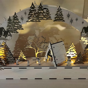 May include: A wooden Christmas village scene with a white cottage, a tree, and a snowman. The scene is illuminated with warm white LED lights.