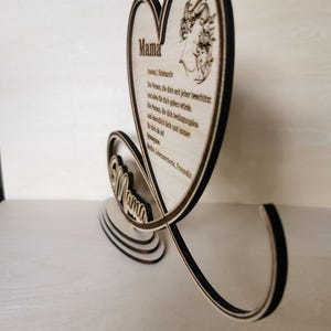 May include: Wooden heart-shaped sign with the word "Mama" in a decorative font. The sign features a small illustration of a mother and child inside the heart. The sign also includes a poem about mothers in German.