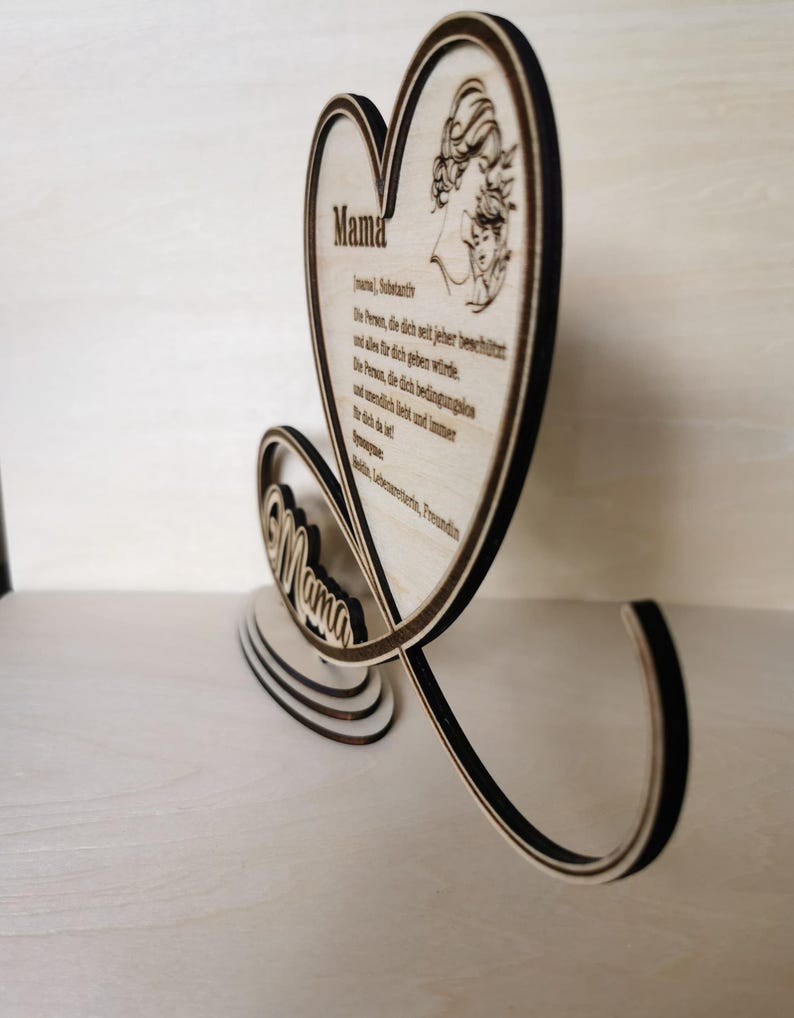 May include: Wooden heart-shaped sign with the word "Mama" in a decorative font. The sign features a small illustration of a mother and child inside the heart. The sign also includes a poem about mothers in German.