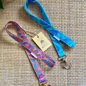May include: Two colourful lanyards with a repeating pattern of circles and dots. The lanyard on the left is pink and orange with blue accents. The lanyard on the right is blue with white accents. Both lanyards have a metal clasp and a tag that reads "Gara Gay" Lanyard Handmade in Australia by Tamara Loutit.