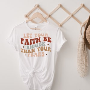 May include: White t-shirt with a motivational quote in a retro font: "Let your faith be bigger than your fears". The text is in shades of red, orange, pink, and blue.