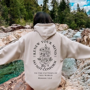 May include: A beige hoodie with a black floral design and the text "Renew Your Mind || Do Not Conform" in a circle. The text "To the pattern of this world Romans 12:2" is below the circle.
