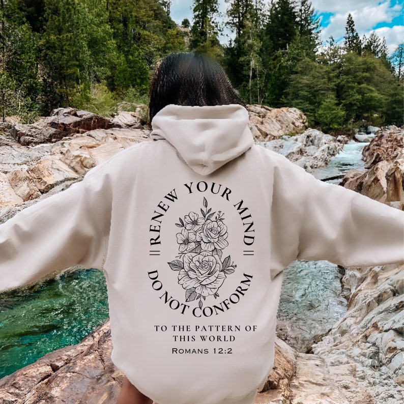 May include: A beige hoodie with a black floral design and the text "Renew Your Mind || Do Not Conform" in a circle. The text "To the pattern of this world Romans 12:2" is below the circle.