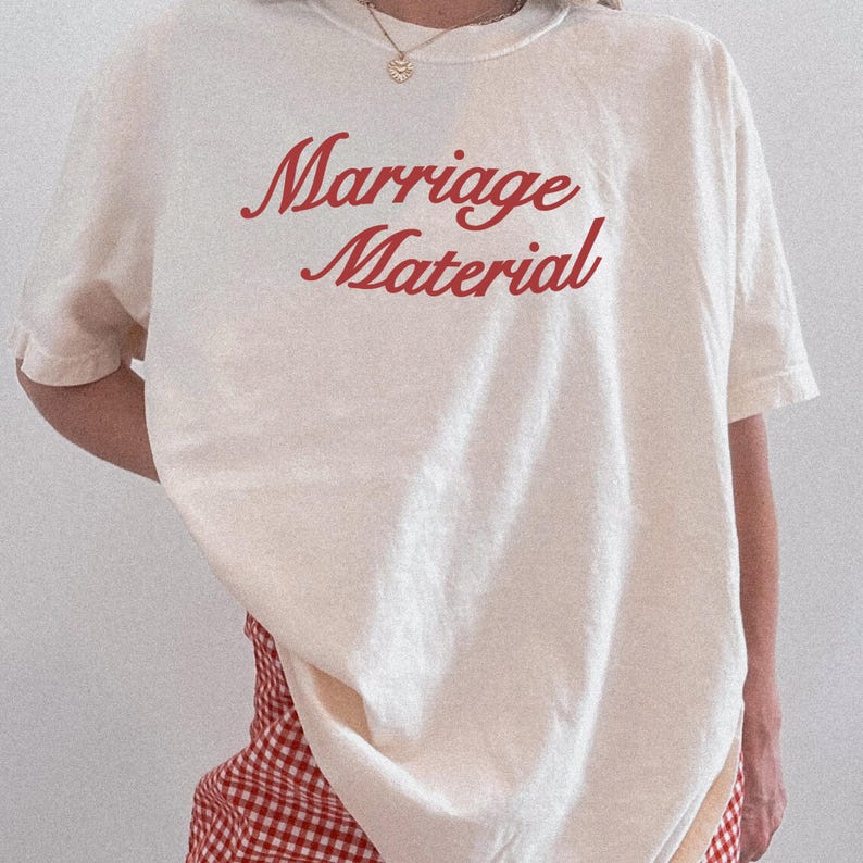 Puede incluir: Camiseta color crema con las palabras "Marriage Material" en escritura roja. La camiseta tiene mangas cortas y un corte relajado. La persona lleva un collar dorado y una falda a cuadros rojos y blancos.