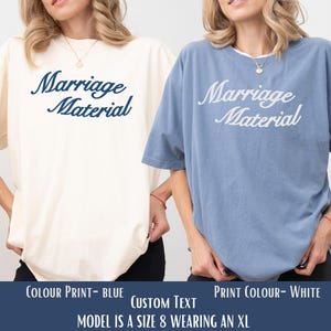 Puede incluir: Dos camisetas, una crema y otra azul, cada una con el texto "Marriage Material" en cursiva. La camiseta crema tiene texto azul, y la azul, texto blanco. La imagen muestra "COLOUR PRINT- BLUE" y "PRINT COLOUR- WHITE".