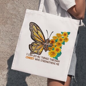 Pu&ograve; includere: Borsa di tela bianca con una farfalla gialla e girasoli. La farfalla ha una croce sulle ali. Il testo sulla borsa recita "Posso fare ogni cosa in Cristo che mi fortifica."