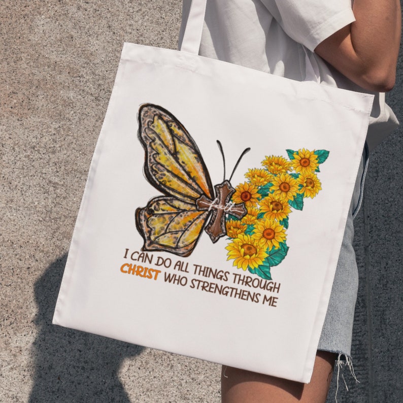 Pu&ograve; includere: Borsa di tela bianca con una farfalla gialla e girasoli. La farfalla ha una croce sulle ali. Il testo sulla borsa recita "Posso fare ogni cosa in Cristo che mi fortifica."