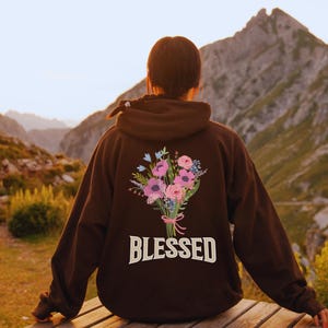 May include: A brown hoodie with a floral bouquet design on the back, featuring pink and purple flowers with blue accents, tied with a pink ribbon. The word "BLESSED" is printed below the bouquet in white. The person is sitting on a wooden bench with a mountain backdrop.