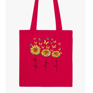 Pu&ograve; includere: Una borsa tote rosa brillante con un design di tre girasoli con farfalle che volano intorno. I girasoli sono etichettati con "fede", "speranza" e "amore".