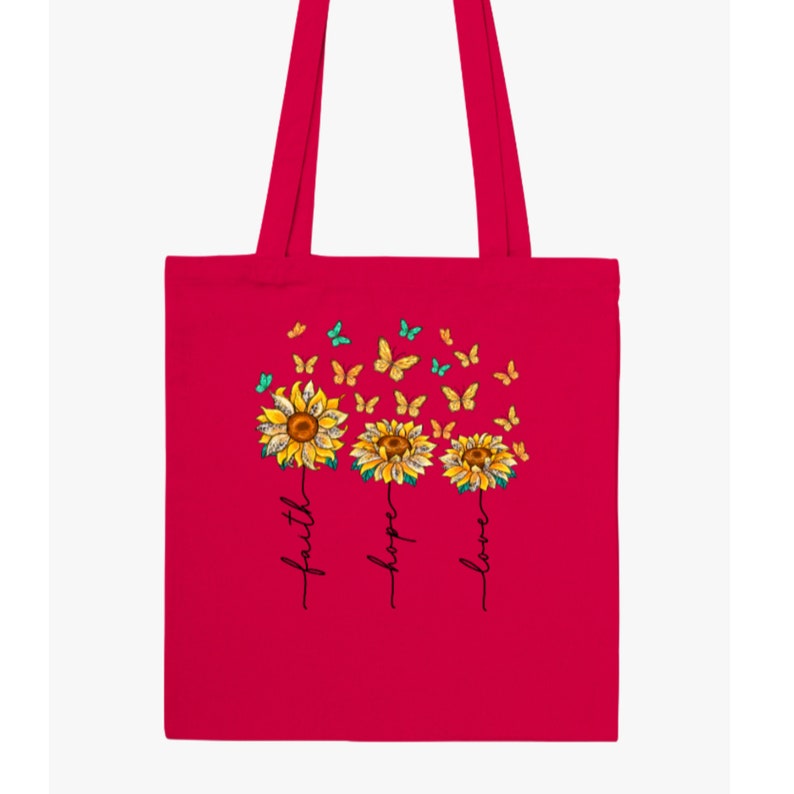 Pu&ograve; includere: Una borsa tote rosa brillante con un design di tre girasoli con farfalle che volano intorno. I girasoli sono etichettati con "fede", "speranza" e "amore".