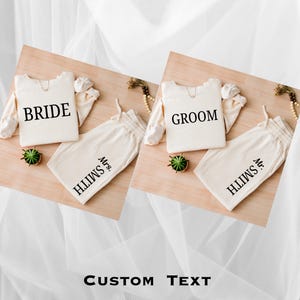 May include: Ivory-coloured matching pyjama sets for a wedding. Each set includes a long-sleeved top with "BRIDE" or "GROOM" printed in black, and matching trousers with "Mrs. Smith" or "Mr. Smith" printed on them. The sets are displayed on a wooden surface.