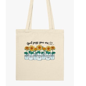 Pu&ograve; includere: Una borsa tote beige con il testo "God says you are" e un simbolo di cuore. La borsa presenta una fila di barattoli di vetro con girasoli e le parole "Lovely", "Terrific", "Strong", "Precious", "Chosen", "Special" e "Unique" scritte su di essi.