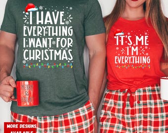 May include: A couple wearing matching Christmas themed t-shirts. The man's shirt says "I have everything I want for Christmas" and the woman's shirt says "It's me I'm everything". Both shirts feature a red Santa hat, snowflakes, and Christmas lights.