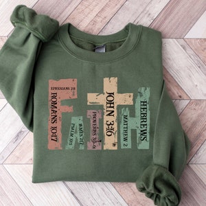 May include: An olive green sweatshirt with the word "FAITH" formed by crosses in various colours. The crosses feature Bible verses such as John 3:16, Romans 10:17, and Hebrews. The sweatshirt is folded on a wooden surface.