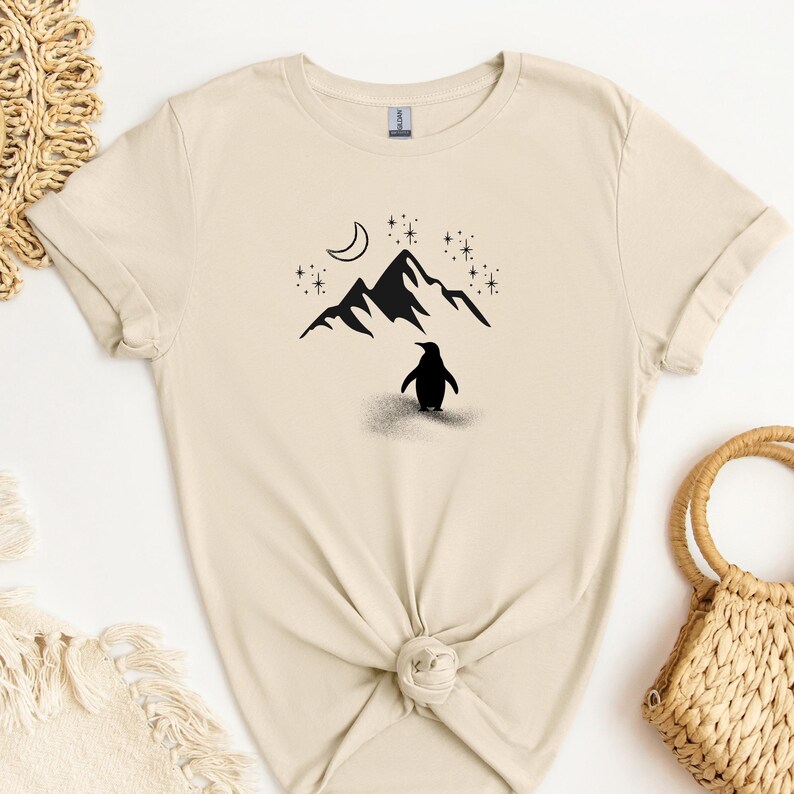 Op de afbeelding: Cr&egrave;mekleurig T-shirt met een zwarte afbeelding van een pingu&iuml;n voor een bergketen, een maan en sterren. Het shirt is in de taille geknoopt.