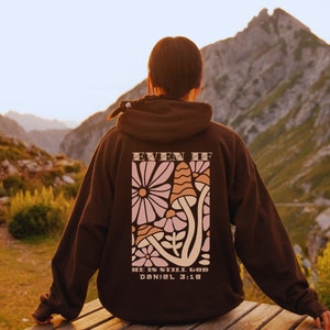 Op de afbeelding: Een bruine hoodie met een bloemen- en paddestoelontwerp op de rug. Het ontwerp bevat de tekst "EVEN IF" bovenaan en "HE IS STILL GOD DANIEL 3:18" onderaan. De hoodie wordt gedragen door een persoon die buiten zit.