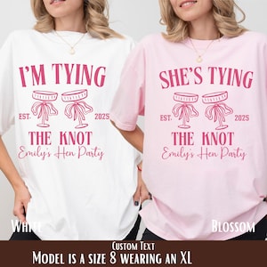 Puede incluir: Dos camisetas, una blanca y otra rosa, con el texto "I'M TYING THE KNOT Emily's Hen Party" y "SHE'S TYING THE KNOT Emily's Hen Party" en rosa. Las camisetas también tienen un gráfico de dos taburetes con lazos y el año 2025.