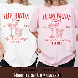 Op de afbeelding: Twee T-shirts, één witte en één roze, met bijpassende rode tekst en afbeeldingen. Het witte shirt leest "THE BRIDE Club" met "My Hen Party TENERIFE" eronder. Het roze shirt zegt "TEAM BRIDE Club" met "Chloe's Hen Party TENERIFE".