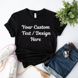 May include: A black t-shirt with the text "Your Custom Text / Design Here" printed in white on the front. The shirt is laid flat on a white surface with a pair of white sneakers, a pair of blue denim shorts, and a wooden bead necklace.