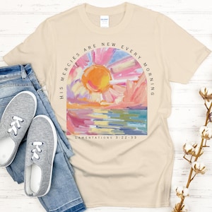 May include: A beige t-shirt with a colorful painting of a sunrise over a body of water. The text "His mercies are new every morning" is written in a semi-circle around the painting. The text "Lamentations 3:22-33" is written below the painting.