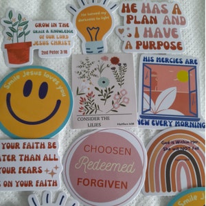 May include: A collection of colorful stickers with inspirational quotes and bible verses. The stickers feature a variety of designs, including flowers, a smiley face, a lightbulb, a rainbow, and a window.