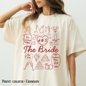 May include: Cream-coloured t-shirt with a crimson graphic design. The design includes the text "The Bride", "It's Giving Wifey", and "Future Mrs. Goy", along with wedding-themed illustrations like doves, rings, and a cake. The print colour is crimson.
