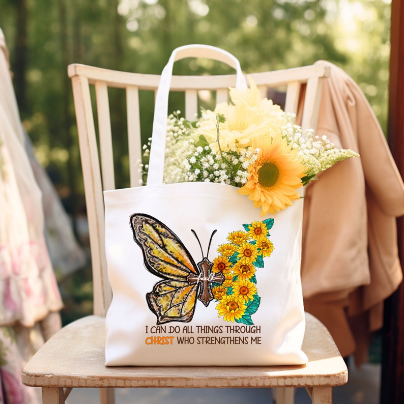 Pu&ograve; includere: Borsa di tela bianca con una farfalla e girasoli. La farfalla ha una croce sulle ali. Il testo sulla borsa recita "Posso fare ogni cosa in Cristo che mi fortifica."