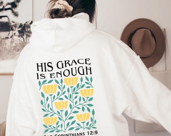 Puede incluir: Sudadera con capucha blanca con un diseño floral y el texto "HIS GRACE IS ENOUGH" y "2ND CORINTHIANS 12:9" impreso en la parte posterior.