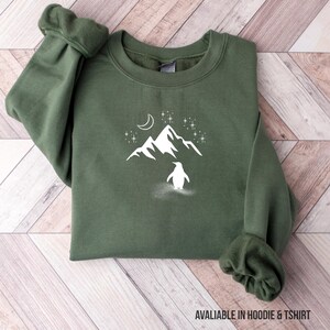 Op de afbeelding: Groen sweatshirt met een witte afbeelding van een pingu&iuml;n, berg, maan en sterren. De tekst "AVALIABLE IN HOODIE & TSHIRT" staat onderaan. Het sweatshirt is op een houten oppervlak gevouwen.