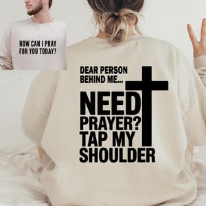 Pode incluir: Camisola bege com o texto "HOW CAN I PRAY FOR YOU TODAY?" na frente e "DEAR PERSON BEHIND ME... NEED PRAYER? TAP MY SHOULDER" com uma cruz nas costas. A camisola é de cor neutra.