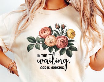 May include: Cream-colored sweatshirt featuring a floral design with pink, peach, and yellow roses, green leaves, and the text "IN THE waiting, GOD IS WORKING" in black script. The sweatshirt has a crew neck.