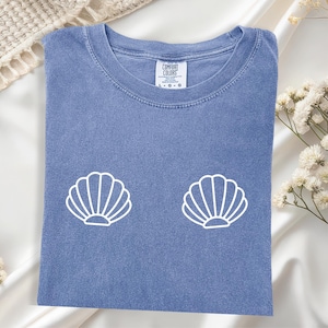 May include: A blue t-shirt with two white seashell graphics. The shirt has a crew neck and short sleeves. The tag reads "COMFORT COLORS" and the size is L-G-G.