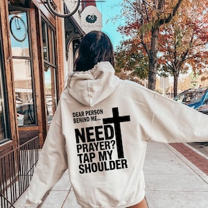 May include: Cream-colored hoodie with black text and a cross graphic. The text reads "DEAR PERSON BEHIND ME... NEED PRAYER? TAP MY SHOULDER." The hoodie is being worn outdoors.