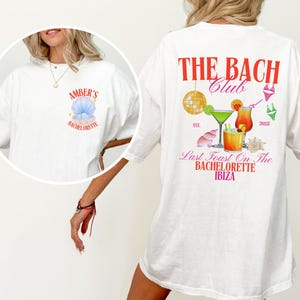 May include: White t-shirt with a front graphic of a blue seashell and the text "AMBER'S BACHELORETTE". The back features the text "THE BACH Club" with cocktail illustrations, a disco ball, and the text "Last Toast On The BACHELORETTE IBIZA".