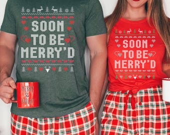 Op de afbeelding: Twee mensen dragen bijpassende kerstshirts en een geruite pyjamabroek. De shirts zijn groen en rood, met de tekst "SOON TO BE MERRY'D" in het wit. Eén persoon houdt een rode mok vast met de tekst "MERRY & BRIGHT".