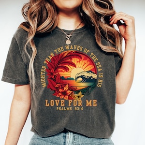 Könnte beinhalten: Ein dunkelgraues T-Shirt mit einer Grafik im Vintage-Stil eines Sonnenuntergangs über dem Meer mit brechenden Wellen. Die Grafik ist von einem Kreis mit dem Text "MIGHTER THAN THE WAVES OF THE SEA IS HIS LOVE FOR ME" und "PSALMS 93:4" umgeben.
