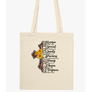 Pu&ograve; includere: Una borsa tote beige con un design a croce marrone e il testo "GOD SAYS YOU ARE" e un elenco di parole e versetti biblici che descrivono attributi positivi. La croce &egrave; decorata con girasoli.