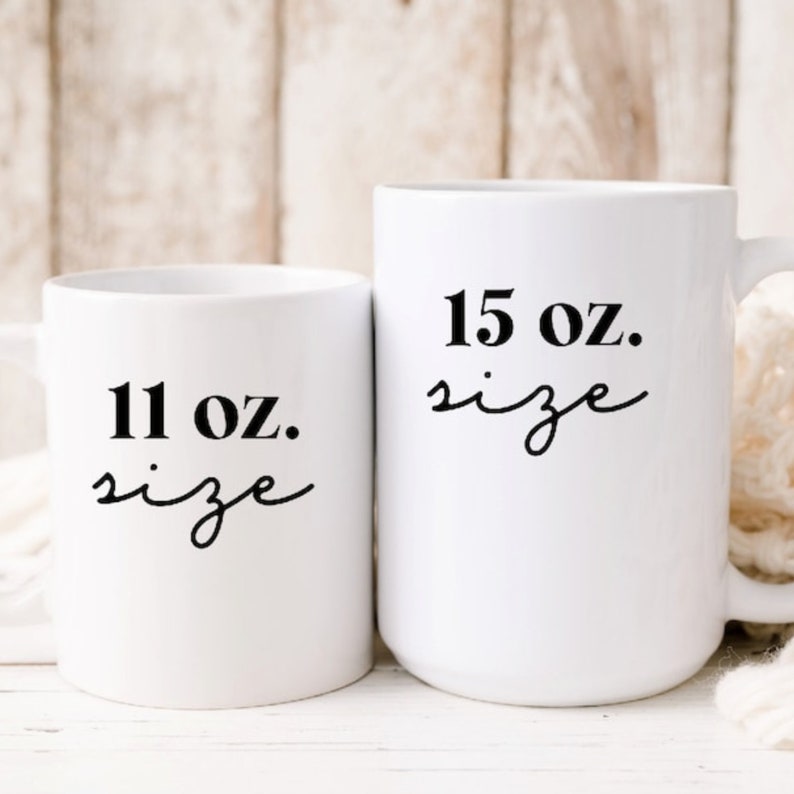 Op de afbeelding: Twee witte keramische mokken met zwarte tekst. De kleinere mok heeft de tekst "11 oz. size" en de grotere mok heeft de tekst "15 oz. size".