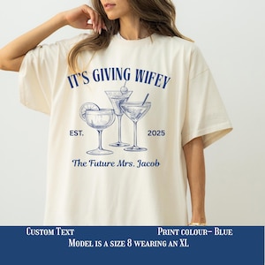 May include: A cream-coloured t-shirt with the text "IT'S GIVING WIFEY" and "The Future Mrs. Jacob" in blue. The shirt features a graphic of three cocktail glasses. The year "2025" and "EST." are also printed on the shirt.