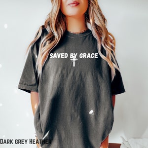 May include: A dark grey heather t-shirt with the text "SAVED BY GRACE" and a white cross printed on the front.